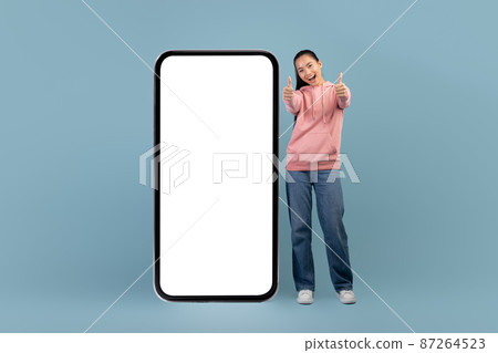 Cheerful asian woman posing with big smartphone, showing thumbs up Cheerful asian woman posing with big smartphone, showing thumbs up 87264523