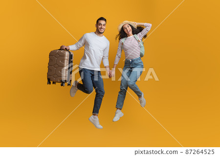 Active Travel Concept. Joyful Arab Couple Jumping With Suitcase On Yellow Background Active Travel Concept. Joyful Arab Couple Jumping With Suitcase On Yellow Background 87264525