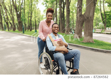 Happy young black guy in wheelchair carrying paper bag with products, going shopping with his girlfriend, outdoors 87264562