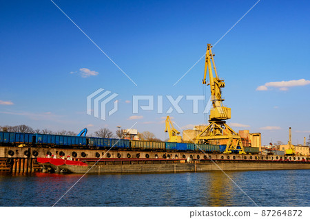 River port crane loading open-top gondola cars on sunny day. Empty river drag boats or barges moored by pier, Empty cars ready for loading. River port crane loading open-top gondola cars on sunny day. Empty river drag boats or barges moored by pier, Empty cars ready for loading. 87264872