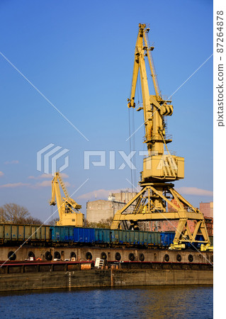 River port crane loading open-top gondola cars on sunny day. Empty river drag boats or barges moored by pier, Empty cars ready for loading. River port crane loading open-top gondola cars on sunny day. Empty river drag boats or barges moored by pier, Empty cars ready for loading. 87264878