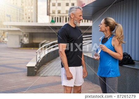 Lovely sportive middle aged couple, man and woman in sportswear smiling at each other, standing together outdoors ready for workout Lovely sportive middle aged couple, man and woman in sportswear smiling at each other, standing together outdoors ready for workout 87265279