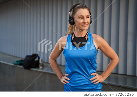Portrait of attractive middle aged sportswoman in sportswear and headphones smiling, listening to music or podcast while standing outdoors, ready for workout Portrait of attractive middle aged sportswoman in sportswear and headphones smiling, listening to music or podcast while standing outdoors, ready for workout 87265280