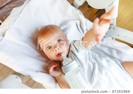 Cute little newborn girl drinking milk from bottle and looking at camera on white background. Infant baby sucking eating milk nutrition lying down on crib bed at home. Motherhood happy child concept Cute little newborn girl drinking milk from bottle and looking at camera on white background. Infant baby sucking eating milk nutrition lying down on crib bed at home. Motherhood happy child concept 87265969