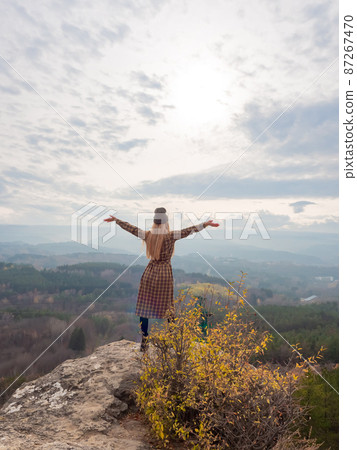 A long haired woman in a dress stands on the edge of a cliff with her arms outstretched and looking into a wooded mountain autumn valley under a cloudy sky 87267470