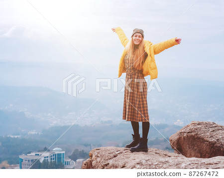 A smiling woman in a plaid dress stands on the edge of a cliff in a smoky hilly valley of the Kislovodsk national park 87267472