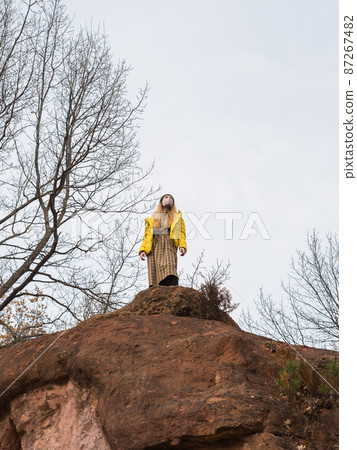 A woman in a face mask stands on top of a cliff near bare trees against a cloudy sky 87267482