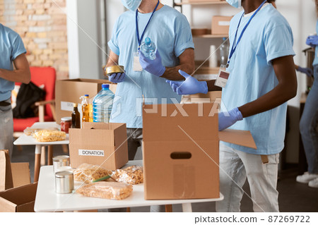 Cropped shot of mutiethnic volunteers in blue uniform, protective gloves separating donations stuffs together, sorting items in cardboard boxes while standing indoors Cropped shot of mutiethnic volunteers in blue uniform, protective gloves separating donations stuffs together, sorting items in cardboard boxes while standing indoors 87269722