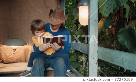 Man in a cowboy hat reads a paper book to a small child sitting on the porch of a village house. The concept of reading paper books, studying and hobbies. Old, retro decor, burlap background Man in a cowboy hat reads a paper book to a small child sitting on the porch of a village house. The concept of reading paper books, studying and hobbies. Old, retro decor, burlap background 87270199