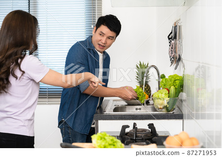 stressed couple conflict in their kitchen, angry woman pointing hand to man for washing vegetables 87271953