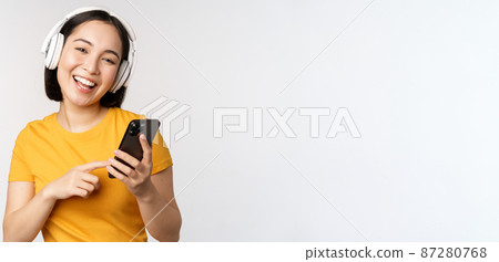 Cute japanese girl in headphones, looking at mobile phone and smiling, using music app on smartphone, standing against white background 87280768