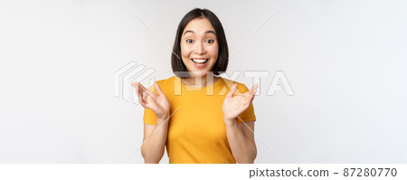 Close up portrait of asian woman looking surprised, wow face, staring impressed at camera, standing over white background in yellow t-shirt 87280770