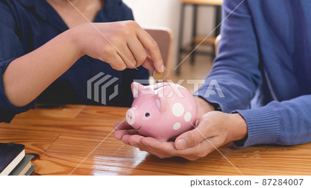 saving concept the female who wear long sleeved shirt trying to drop a coin into a pigging bank while the guy in lighter blue sweater carrying it for her 87284007