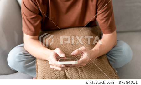 Technology Concept The man sitting next to a pot of plant, putting a small pillow on his lap, and checking something on his smartphone Technology Concept The man sitting next to a pot of plant, putting a small pillow on his lap, and checking something on his smartphone 87284011
