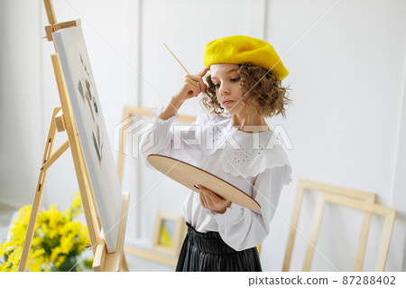 Portrait of cute thoughtful little girl artist in yellow beret who paints picture in studio while holds color pallete 87288402