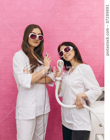 Woman holding a working part of the epilator in hands. Cosmetologist doctor with needle of electro epilation. Beauty industry, treatment at salon, electrolysis. Two girls standing on pink background Woman holding a working part of the epilator in hands. Cosmetologist doctor with needle of electro epilation. Beauty industry, treatment at salon, electrolysis. Two girls standing on pink background 87289891