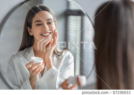 Cosmetics Concept. Young Woman Looking At Mirror And Applying Face Cream Cosmetics Concept. Young Woman Looking At Mirror And Applying Face Cream 87290031