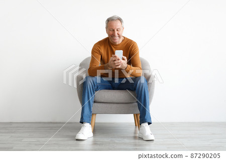 Portrait of smiling mature man using smartphone sitting on chair Portrait of smiling mature man using smartphone sitting on chair 87290205