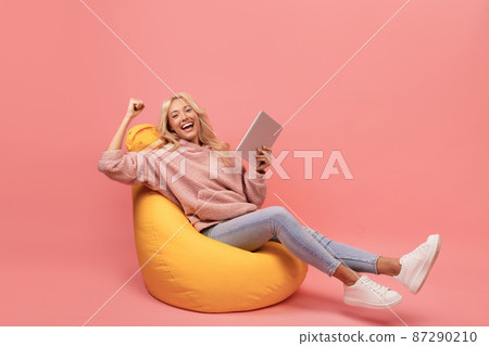 Overjoyed woman holding digital tablet and exclaiming with joy while relaxing in beanbag chair over pink background 87290210