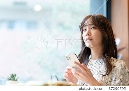 一位年輕女子一邊喝咖啡,一邊在咖啡館的靠窗座位上看著她的智能手機 一位年輕女子一邊喝咖啡,一邊在咖啡館的靠窗座位上看著她的智能手機 87290795