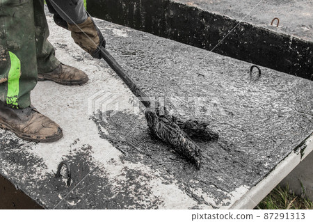 A construction worker is engaged in dirty manual work to repair and apply black bitumen waterproofing tar to the concrete surface of the structure A construction worker is engaged in dirty manual work to repair and apply black bitumen waterproofing tar to the concrete surface of the structure 87291313