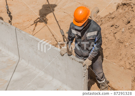 An industrial worker in overalls and a protective helmet attaches a heavy iron hook lifting mechanism with a steel cable rope to a concrete, cement structure An industrial worker in overalls and a protective helmet attaches a heavy iron hook lifting mechanism with a steel cable rope to a concrete, cement structure 87291328