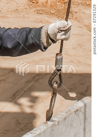 The hand of construction worker male take a heavy iron hook lifting mechanism with a steel cable rope to a concrete, cement structure The hand of construction worker male take a heavy iron hook lifting mechanism with a steel cable rope to a concrete, cement structure 87291329