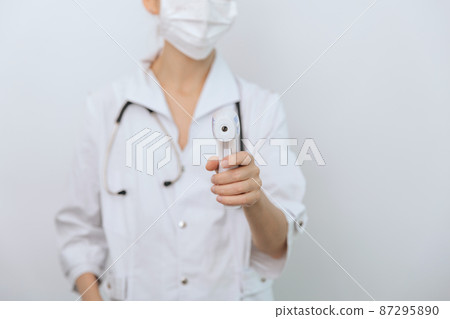 Temperature measurement gun in doctor hands. Close-up shot of doctor wearing protective surgical mask ready to use infrared isometric thermometer gun to check body temperature for virus symptoms. Temperature measurement gun in doctor hands. Close-up shot of doctor wearing protective surgical mask ready to use infrared isometric thermometer gun to check body temperature for virus symptoms. 87295890