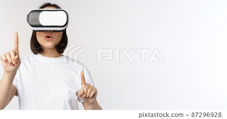 Potrait of asian woman in virtual reality glasses, pointing, choosing smth in VR headset, standing over white background 87296928