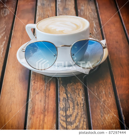 White cup of cappuccino with sunglasses on a saucer in front of it stands on a wooden surface White cup of cappuccino with sunglasses on a saucer in front of it stands on a wooden surface 87297444