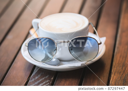 White cup of cappuccino with sunglasses stands on a wooden surface 87297447