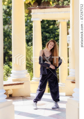 A long-haired woman in wide corduroy pants and a shirt lowered from her shoulders stands in pose with her toes inward against the background of columns 87297552