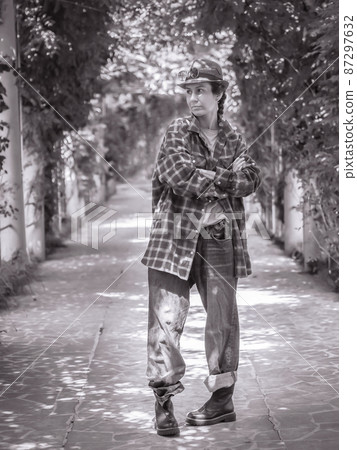 A girl in a hat and casual oversized outfit stands with her legs bent and her arms folded in an alley in the park. Black and white photo. Posterization 87297632