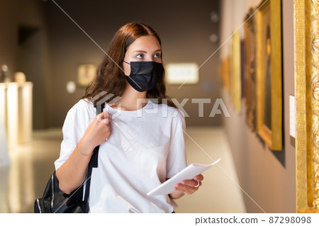 Focused girl in a protective mask looks at the paintings in the museum hall 87298098