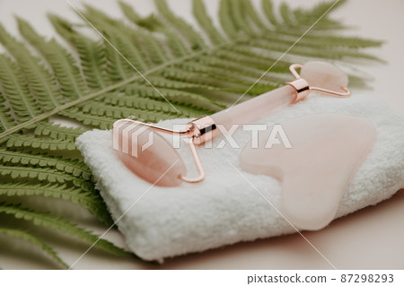 Scin care concept. Roller gua sha massage scraper on a towel with a fern leaf on a pink background Scin care concept. Roller gua sha massage scraper on a towel with a fern leaf on a pink background 87298293