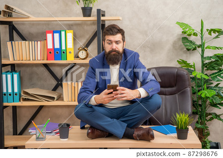 Professional man with puzzled look reading message in smartphone sitting on office desk, sms 87298676