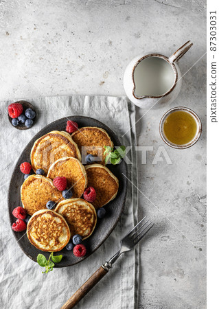 Pancakes in ceramic plate with berries, mint leaves, gravy boats and fork on napkin, top view 87303031