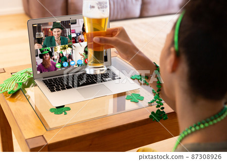 Mixed race woman raising beer glass making st patrick's day video call to friends on laptop at home Mixed race woman raising beer glass making st patrick's day video call to friends on laptop at home 87308926