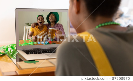 Mixed race woman making st patrick's day video call to friends holding beers on laptop at home 87308970
