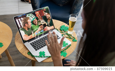 Caucasian woman with beer making st patrick's day video call waving to friends on laptop at bar 87309015