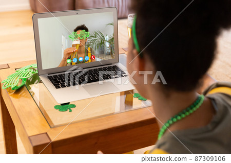 Mixed race woman making st patrick's day video call to friend in costume on laptop at home 87309106