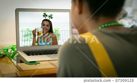 Mixed race woman making st patrick's day video call to female friend holding beer on laptop at home Mixed race woman making st patrick's day video call to female friend holding beer on laptop at home 87309128