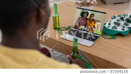 African american man holding beer having st patrick's day video call with friends on laptop at home 87309138