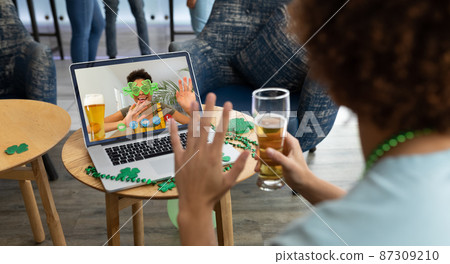 Mixed race man holding beer at bar making st patrick's day video call waving to friend on laptop Mixed race man holding beer at bar making st patrick's day video call waving to friend on laptop 87309210