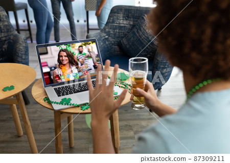Mixed race man holding beer at bar making st patrick's day video call waving to friend on laptop Mixed race man holding beer at bar making st patrick's day video call waving to friend on laptop 87309211