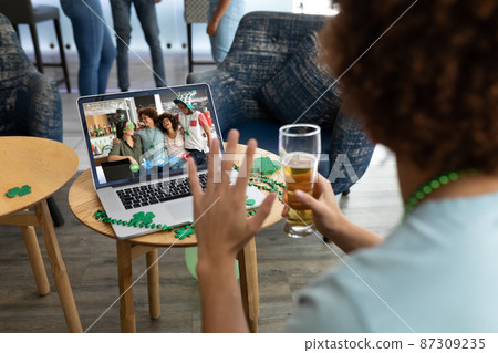Mixed race man holding beer at bar making st patrick's day video call waving to friends on laptop Mixed race man holding beer at bar making st patrick's day video call waving to friends on laptop 87309235