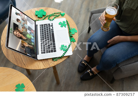 Mixed race woman holding beer at bar making st patrick's day video call with friends on laptop Mixed race woman holding beer at bar making st patrick's day video call with friends on laptop 87309257