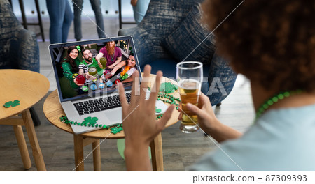 Mixed race man holding beer at bar making st patrick's day video call waving to friends on laptop Mixed race man holding beer at bar making st patrick's day video call waving to friends on laptop 87309393