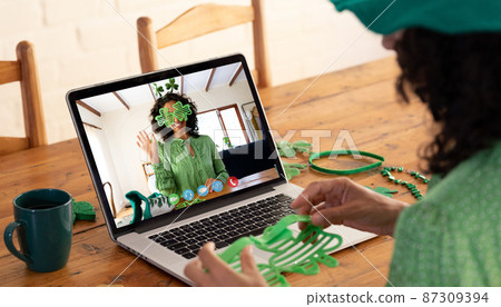 Mixed race woman making st patrick's day video call to waving female friend on laptop at home 87309394