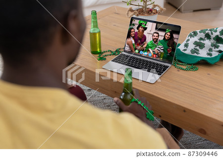 African american man holding beer making st patrick's day video call with friends on laptop at home 87309446